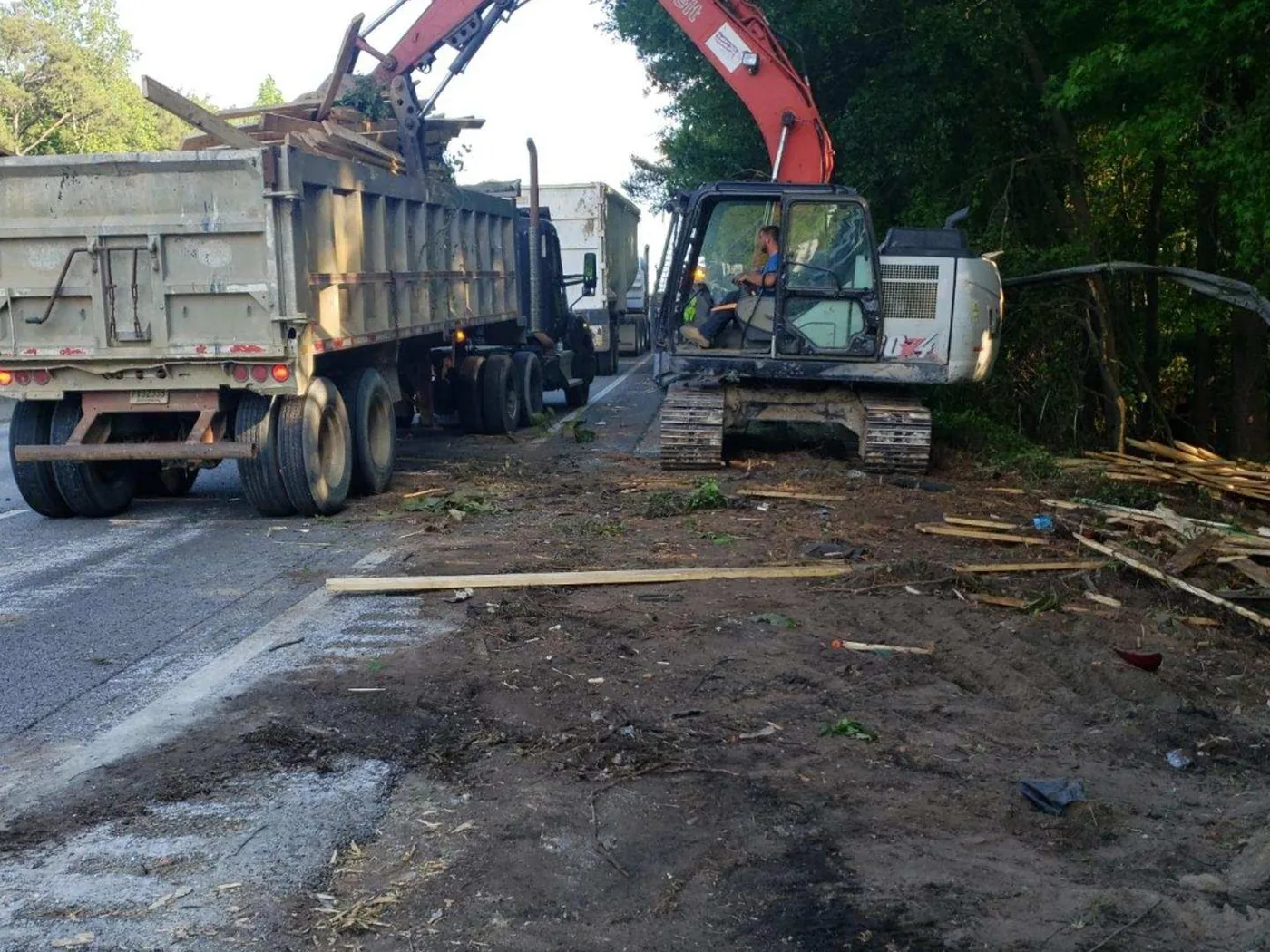 Excavator loading a dump truck at a roadside ditch dig along the tree line