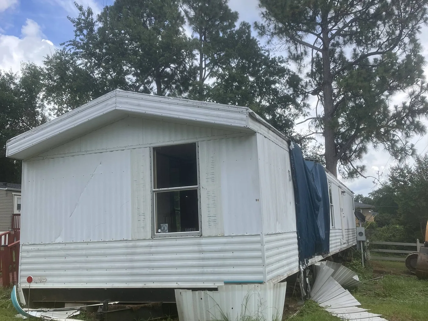 Older single wide mobile home with a tarp over the roof, sitting on its piers before demolition