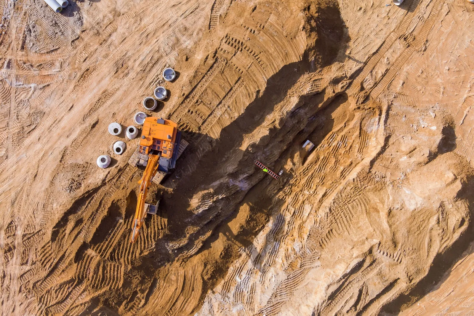 Aerial view of an excavator working a pipe trench on sandy ground, with pipe sections staged beside the cut