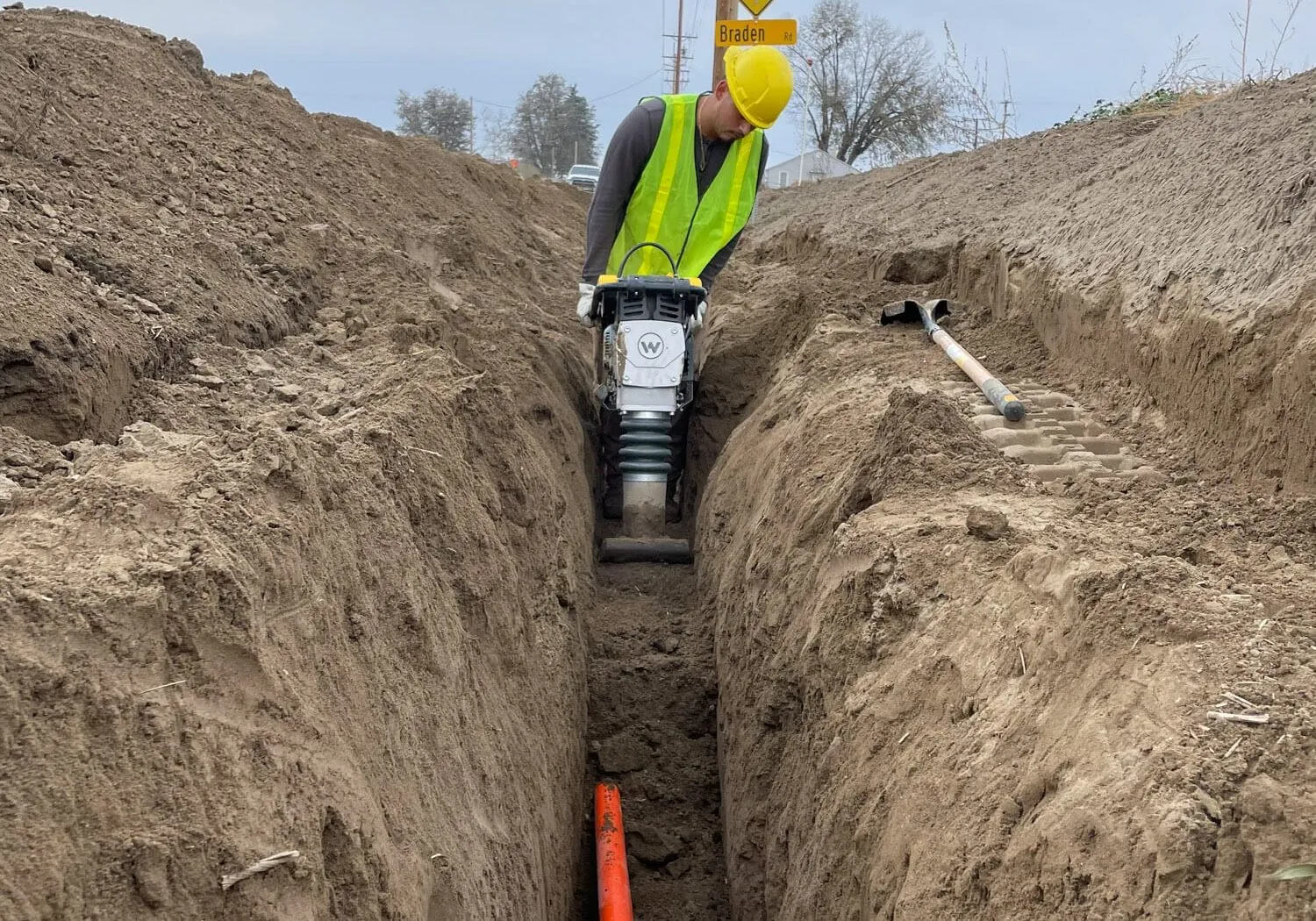 Worker compacting the bottom of a narrow utility trench with a rammer, orange conduit in the trench